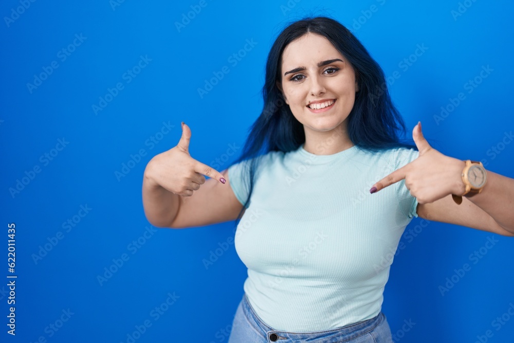 Fototapeta premium Young modern girl with blue hair standing over blue background looking confident with smile on face, pointing oneself with fingers proud and happy.