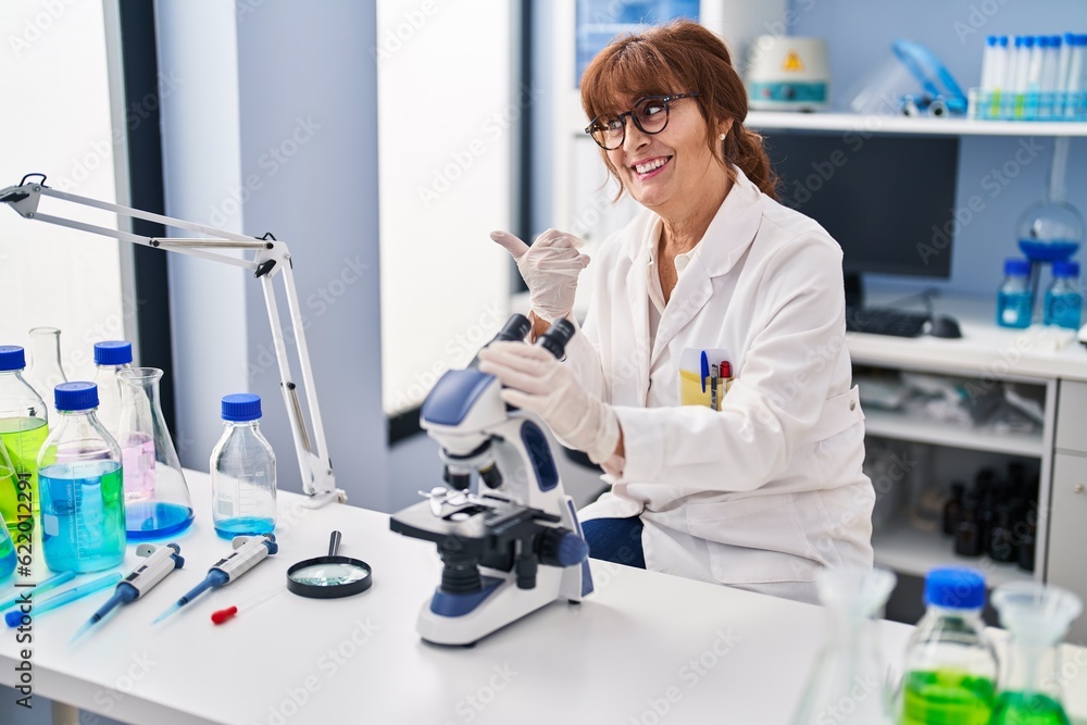 Middle age hispanic woman working at scientist laboratory pointing thumb up to the side smiling happy with open mouth