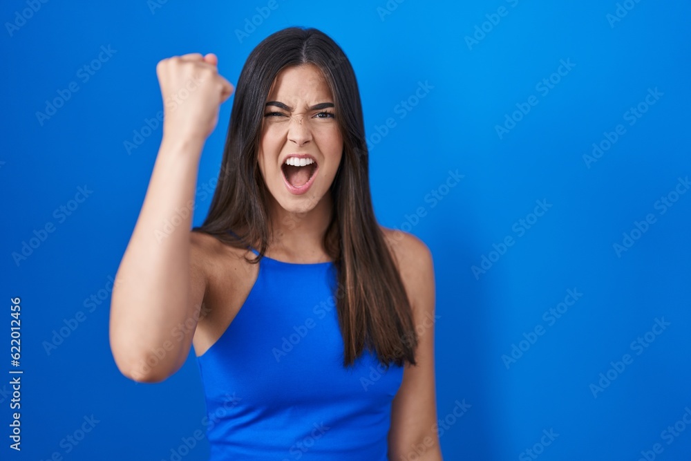 Hispanic woman standing over blue background angry and mad raising fist frustrated and furious while shouting with anger. rage and aggressive concept. Hispanic woman standing over blue background angry and mad raising fist frustrated and furious while shouting with anger. rage and aggressive concept.