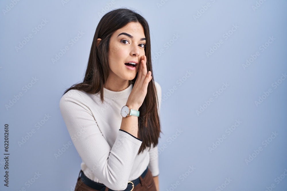 Young brunette woman standing over blue background hand on mouth telling secret rumor, whispering malicious talk conversation - obrazy, fototapety, plakaty 