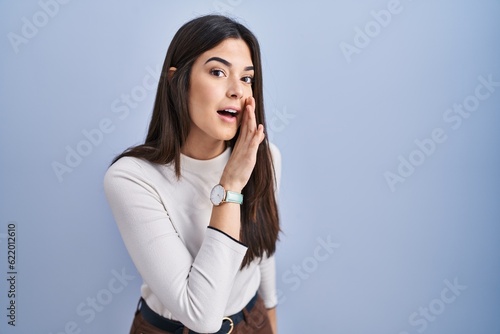 Fototapeta Young brunette woman standing over blue background hand on mouth telling secret