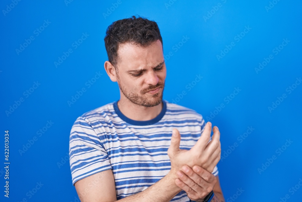 Young hispanic man standing over blue background suffering pain on hands and fingers, arthritis inflammation Young hispanic man standing over blue background suffering pain on hands and fingers, arthritis inflammation