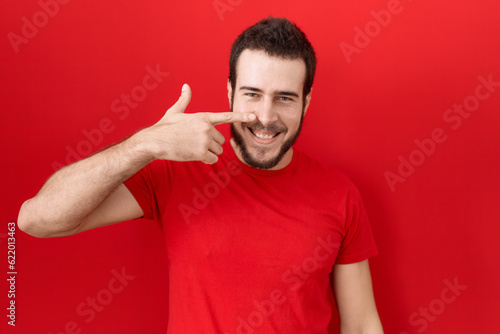 Young hispanic man wearing casual red t shirt pointing with hand finger to face and nose, smiling cheerful. beauty concept