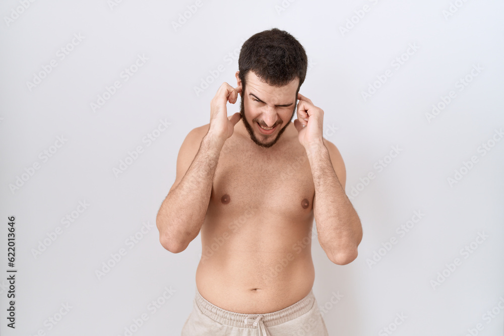 Fototapeta premium Young hispanic man standing shirtless over white background covering ears with fingers with annoyed expression for the noise of loud music. deaf concept.
