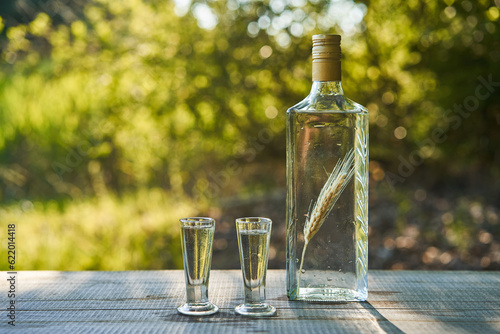Two shots of crystal clear delicious polish vodka in vintage shoot glasses and rustic style bottle with macerated rye ear inside. Served outside on wooden table during sunny summer saturday evening.