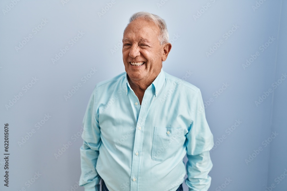 Senior man with grey hair standing over blue background winking looking at the camera with sexy expression, cheerful and happy face. Senior man with grey hair standing over blue background winking looking at the camera with sexy expression, cheerful and happy face.