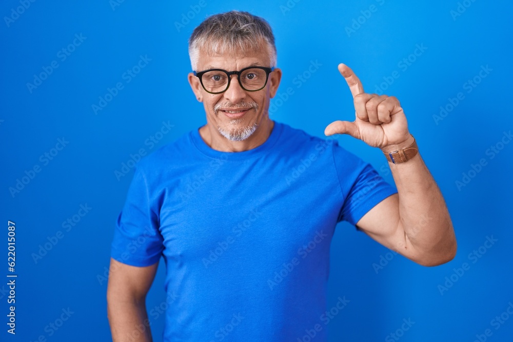 Hispanic man with grey hair standing over blue background smiling and confident gesturing with hand doing small size sign with fingers looking and the camera. measure concept. Hispanic man with grey hair standing over blue background smiling and confident gesturing with hand doing small size sign with fingers looking and the camera. measure concept.