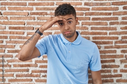 Brazilian young man standing over brick wall worried and stressed about a problem with hand on forehead, nervous and anxious for crisis