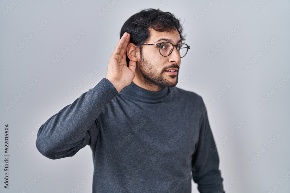 Handsome latin man standing over isolated background smiling with hand over ear listening an hearing to rumor or gossip. deafness concept. Handsome latin man standing over isolated background smiling with hand over ear listening an hearing to rumor or gossip. deafness concept.