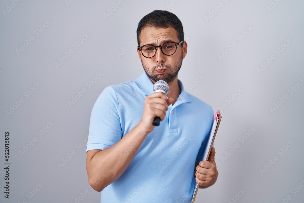 Hispanic man holding reporter microphone and clipboard puffing cheeks with funny face. mouth inflated with air, catching air. Hispanic man holding reporter microphone and clipboard puffing cheeks with funny face. mouth inflated with air, catching air.