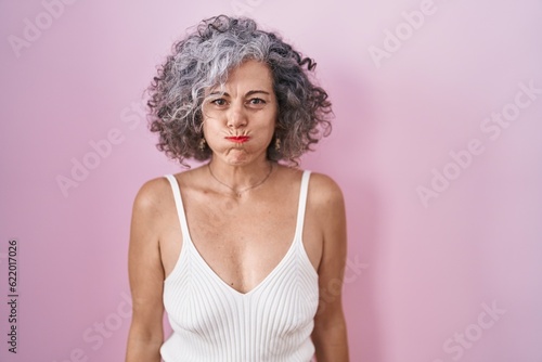 Middle age woman with grey hair standing over pink background puffing cheeks with funny face. mouth inflated with air, crazy expression.