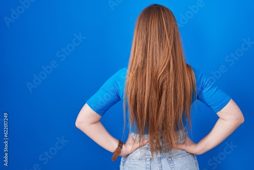 Redhead woman standing over blue background standing backwards looking away with arms on body