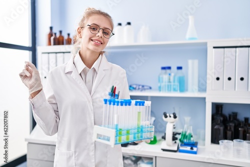 Young caucasian woman working at scientist laboratory holding samples smiling happy pointing with hand and finger to the side