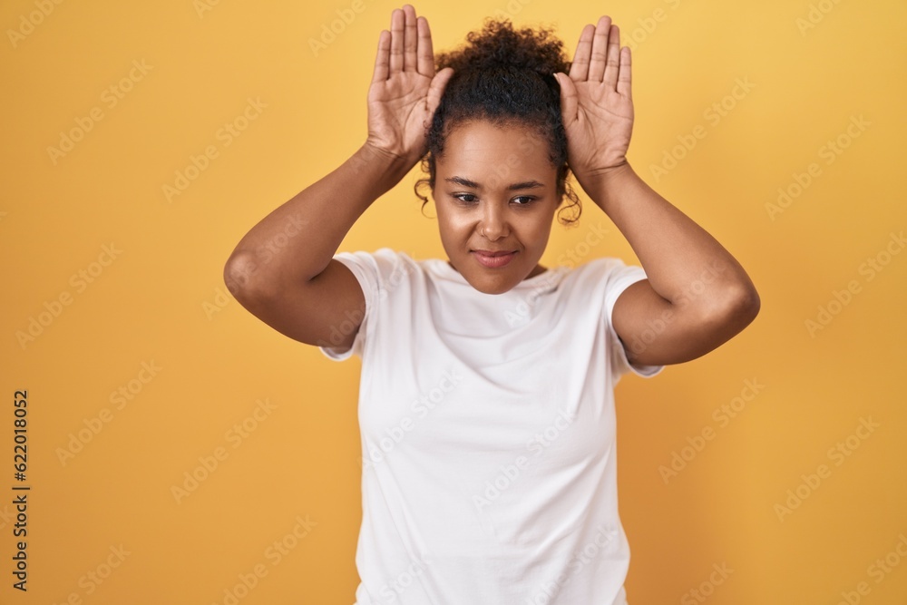 Young hispanic woman with curly hair standing over yellow background doing bunny ears gesture with hands palms looking cynical and skeptical. easter rabbit concept. Young hispanic woman with curly hair standing over yellow background doing bunny ears gesture with hands palms looking cynical and skeptical. easter rabbit concept.