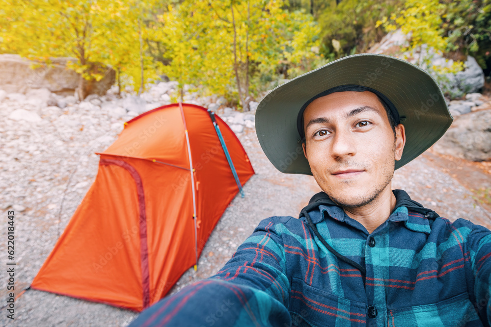 Happy hiker man wearing hat taking selfie photo against red tent in the woods. Solo trail walking and trekking equipment