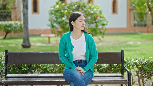 Young chinese woman sitting on bench looking around with serious expression at park
