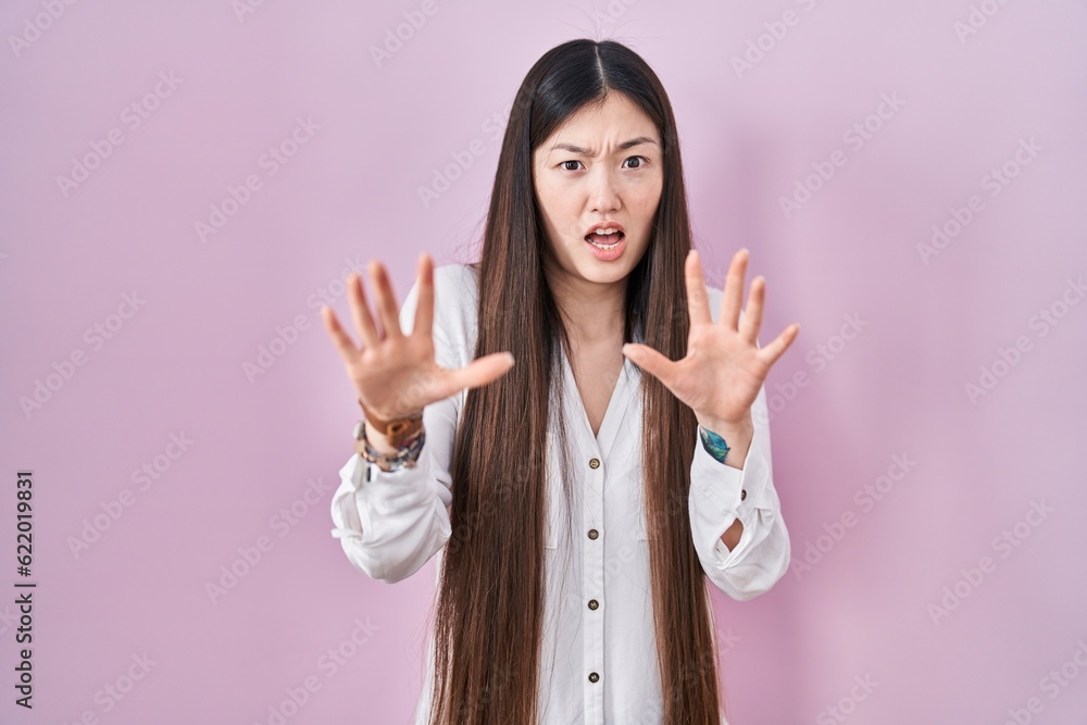 Chinese young woman standing over pink background afraid and terrified with fear expression stop gesture with hands, shouting in shock. panic concept. Chinese young woman standing over pink background afraid and terrified with fear expression stop gesture with hands, shouting in shock. panic concept.