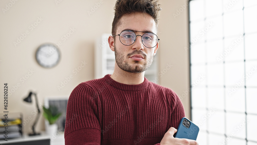 Young hispanic man business worker holding smartphone with serious face at office Young hispanic man business worker holding smartphone with serious face at office