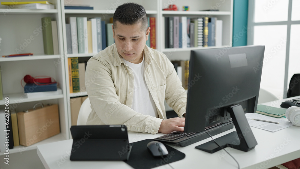 Young hispanic man student using computer and touchpad studying at library university Young hispanic man student using computer and touchpad studying at library university