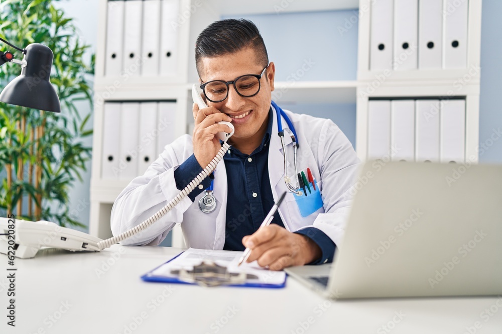 Young latin man doctor talking on telephone writing on document at clinic Young latin man doctor talking on telephone writing on document at clinic