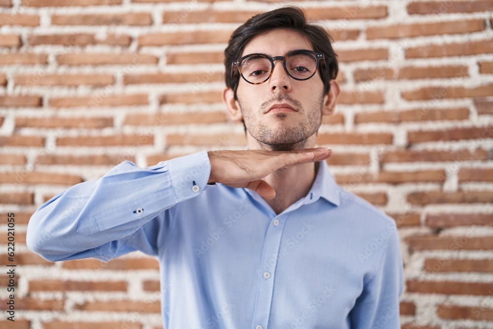 Young hispanic man standing over brick wall background cutting throat with hand as knife, threaten aggression with furious violence Young hispanic man standing over brick wall background cutting throat with hand as knife, threaten aggression with furious violence
