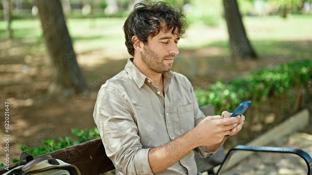 Young hispanic man using smartphone with serious expression sitting on bench at park