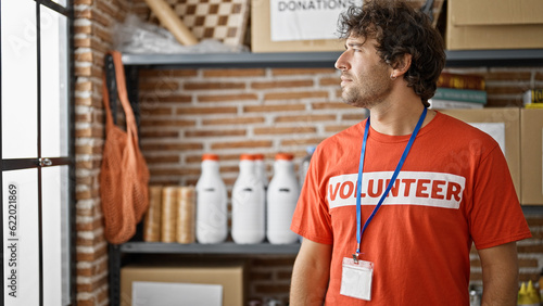 Young hispanic man wearing volunteer uniform looking throw window with serious face at charity center