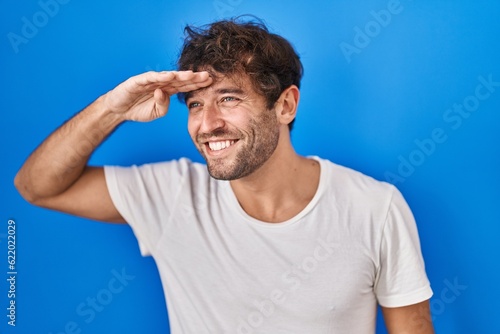 Hispanic young man standing over blue background very happy and smiling looking far away with hand over head. searching concept.