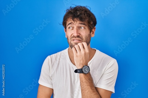 Hispanic young man standing over blue background looking stressed and nervous with hands on mouth biting nails. anxiety problem.