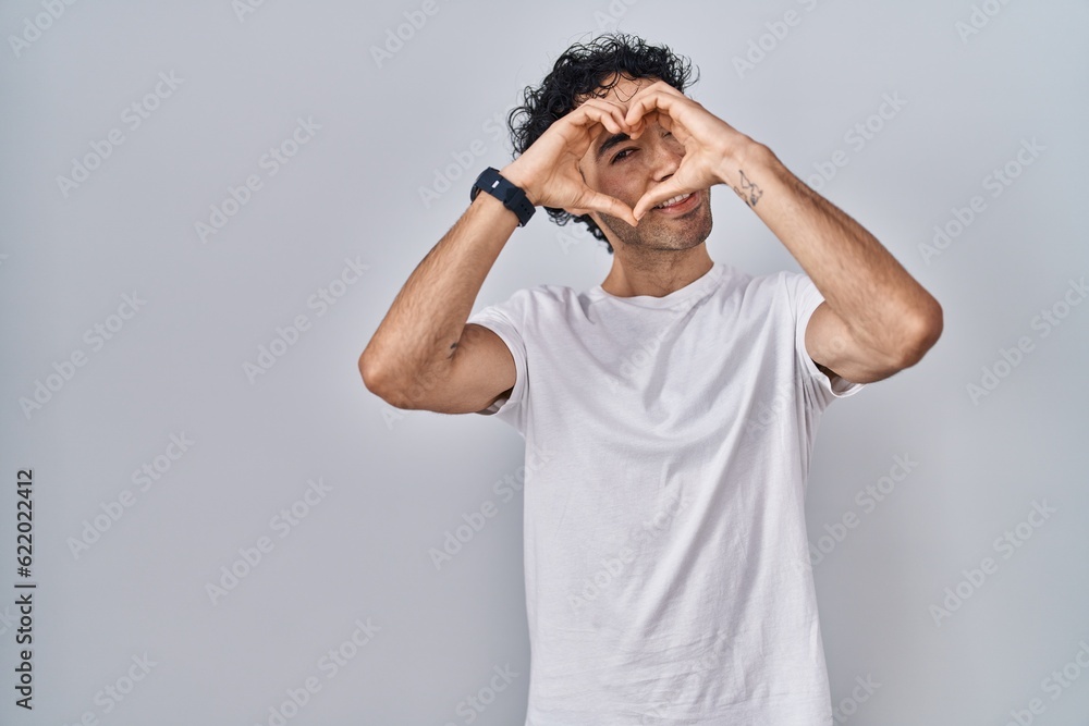 Hispanic man standing over isolated background doing heart shape with hand and fingers smiling looking through sign Hispanic man standing over isolated background doing heart shape with hand and fingers smiling looking through sign