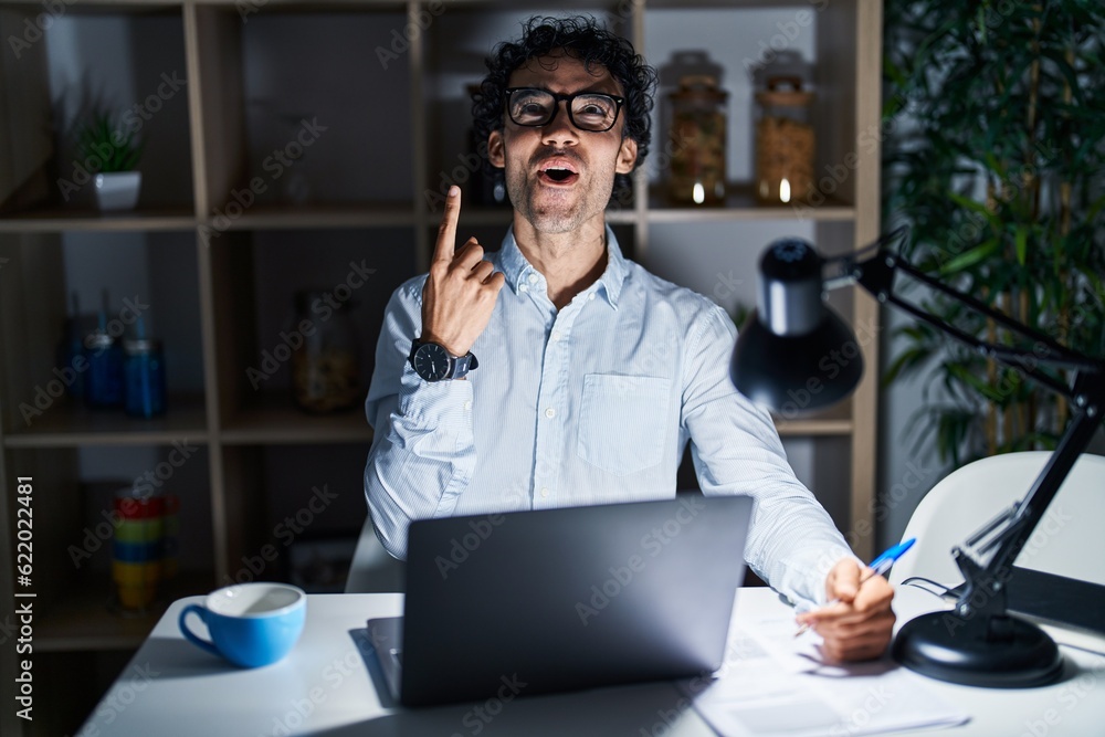 Obraz premium Hispanic man working at the office at night amazed and surprised looking up and pointing with fingers and raised arms.