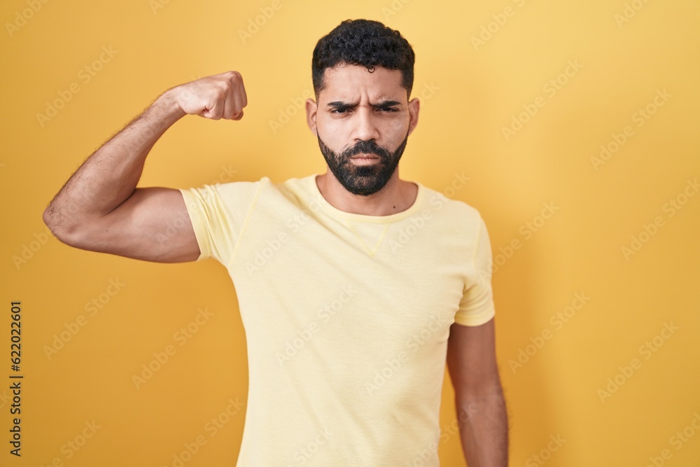Hispanic man with beard standing over yellow background strong person showing arm muscle, confident and proud of power Hispanic man with beard standing over yellow background strong person showing arm muscle, confident and proud of power