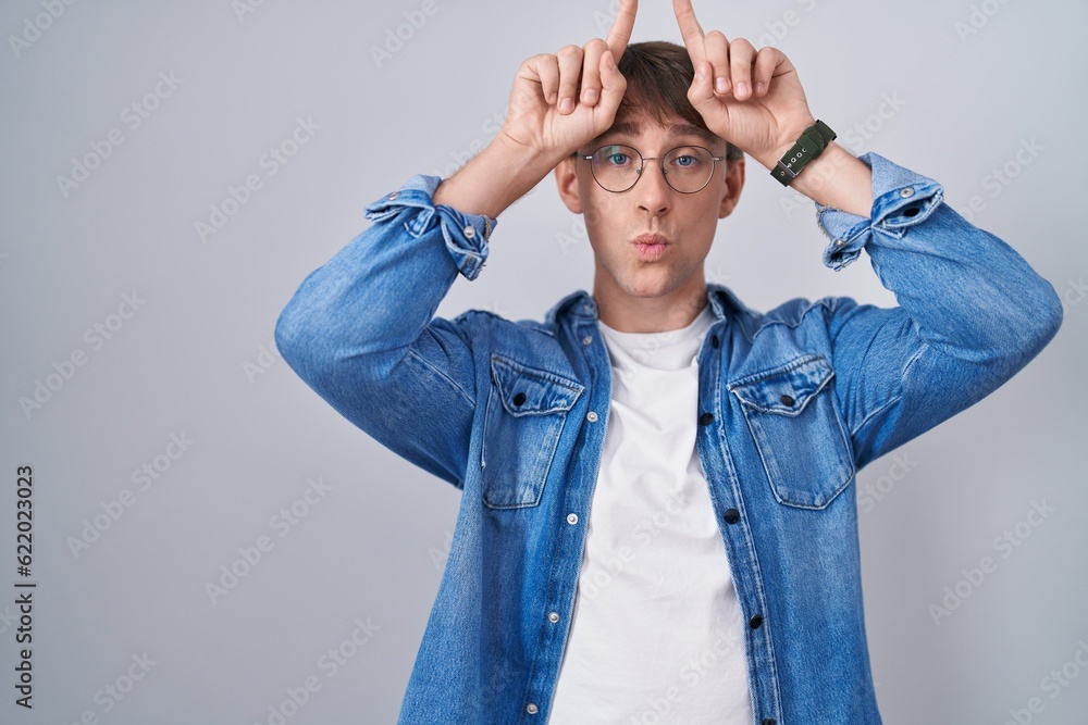 Caucasian blond man standing wearing glasses doing funny gesture with finger over head as bull horns Caucasian blond man standing wearing glasses doing funny gesture with finger over head as bull horns