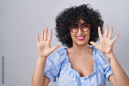 Young brunette woman with curly hair wearing glasses over isolated background showing and pointing up with fingers number ten while smiling confident and happy.