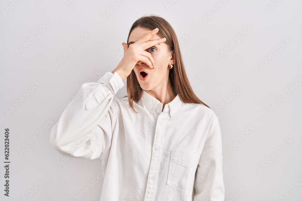 Young caucasian woman standing over isolated background peeking in shock covering face and eyes with hand, looking through fingers with embarrassed expression. Young caucasian woman standing over isolated background peeking in shock covering face and eyes with hand, looking through fingers with embarrassed expression.