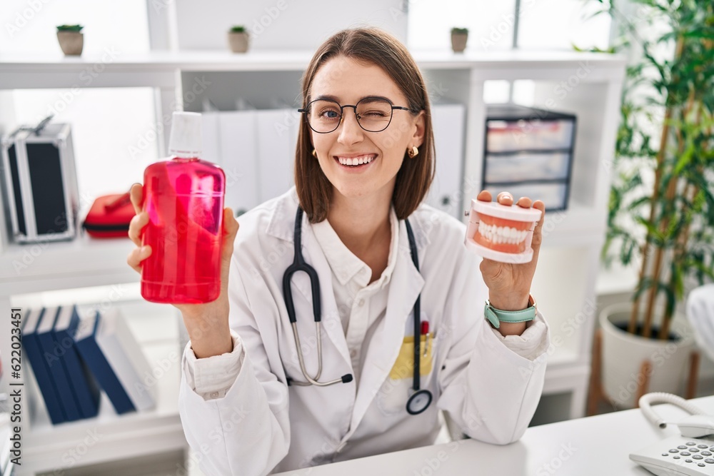 Young caucasian dentist woman holding denture and mouthwash winking looking at the camera with sexy expression, cheerful and happy face. Young caucasian dentist woman holding denture and mouthwash winking looking at the camera with sexy expression, cheerful and happy face.