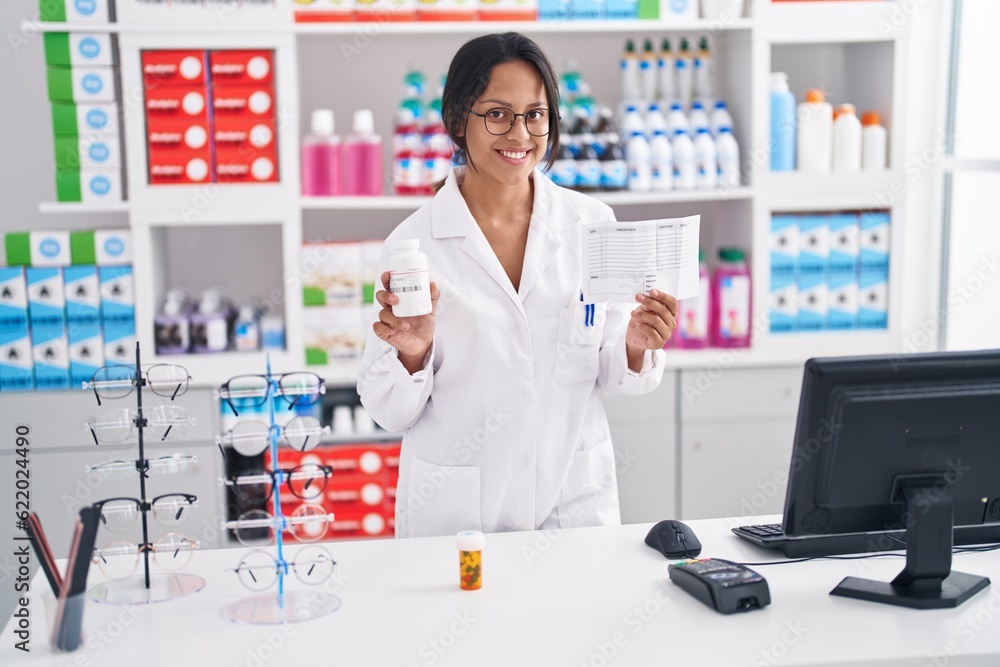 Young hispanic woman pharmacist holding pills bottle and prescription paper at pharmacy