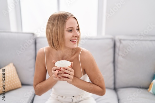 Young blonde woman drinking coffee sitting on sofa at home
