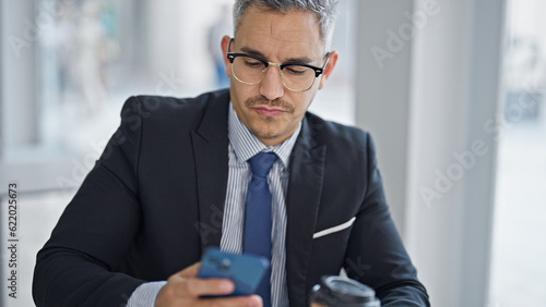 Young hispanic man business worker using smartphone holding coffee at office