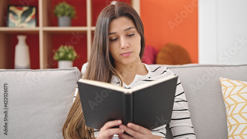 Young beautiful hispanic woman reading book sitting on sofa at home