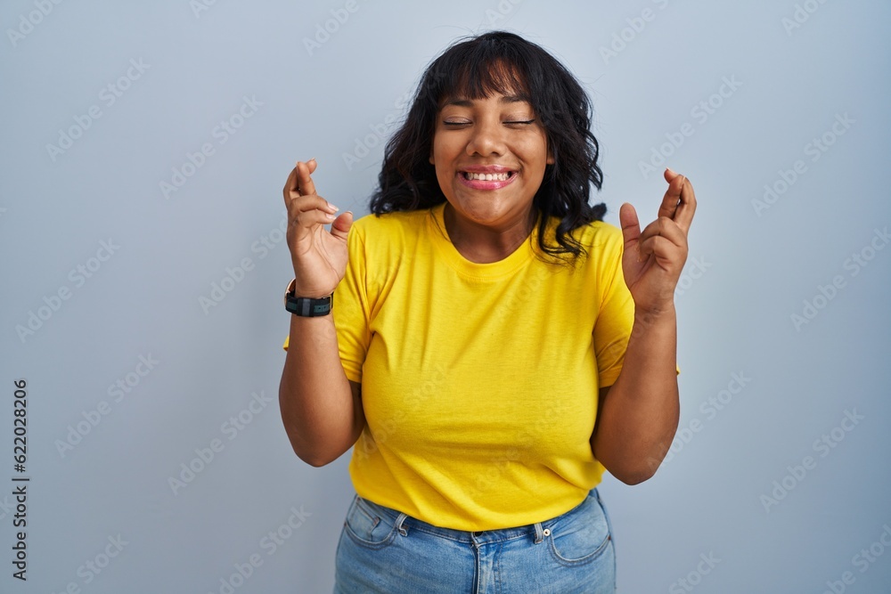 Hispanic woman standing over blue background gesturing finger crossed smiling with hope and eyes closed. luck and superstitious concept. Hispanic woman standing over blue background gesturing finger crossed smiling with hope and eyes closed. luck and superstitious concept.