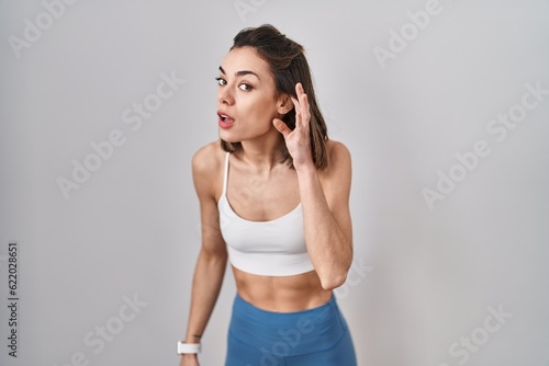 Hispanic woman wearing sportswear over isolated background smiling with hand over ear listening an hearing to rumor or gossip. deafness concept. © Krakenimages.com