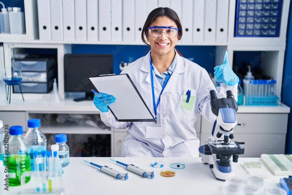 Hispanic young woman working at scientist laboratory doing happy thumbs up gesture with hand. approving expression looking at the camera showing success. Hispanic young woman working at scientist laboratory doing happy thumbs up gesture with hand. approving expression looking at the camera showing success.