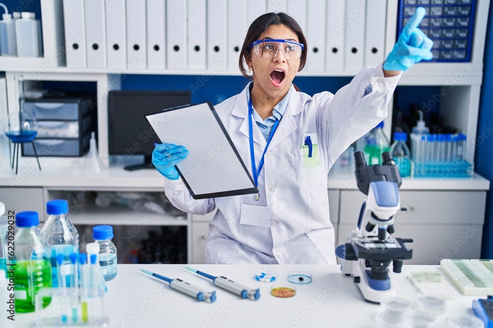 Hispanic young woman working at scientist laboratory pointing with finger surprised ahead, open mouth amazed expression, something on the front Hispanic young woman working at scientist laboratory pointing with finger surprised ahead, open mouth amazed expression, something on the front