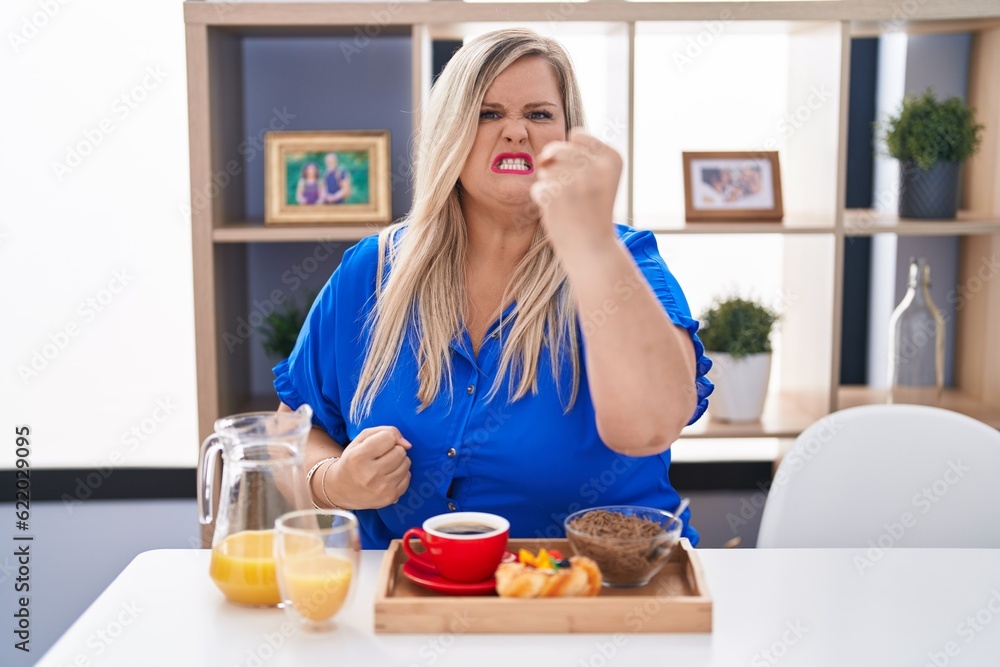 Caucasian plus size woman eating breakfast at home angry and mad raising fist frustrated and furious while shouting with anger. rage and aggressive concept. Caucasian plus size woman eating breakfast at home angry and mad raising fist frustrated and furious while shouting with anger. rage and aggressive concept.