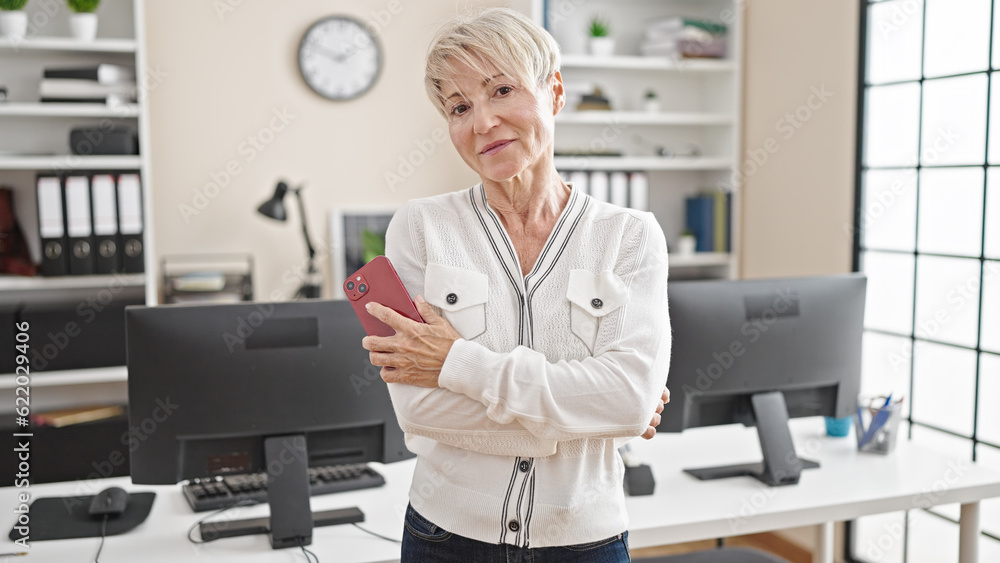 Middle age blonde woman business worker standing with arms crossed gesture holding smartphone at office