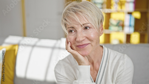 Middle age blonde woman sitting on sofa looking to the side smiling at home