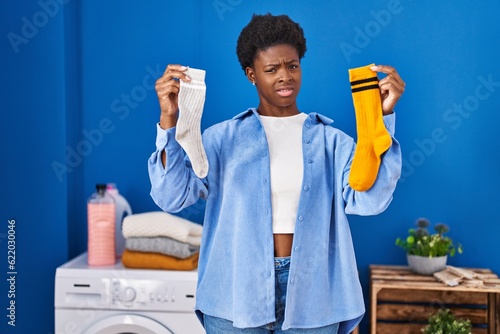 African american woman holding clean andy dirty socks clueless and confused expression. doubt concept. photo