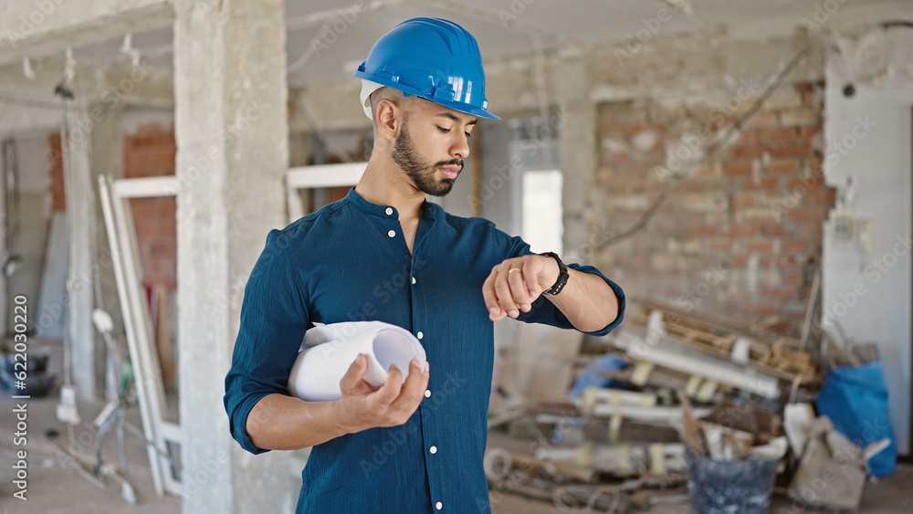 Young hispanic man architect wearing hardhat holding blueprints looking at wrist watch at construction site