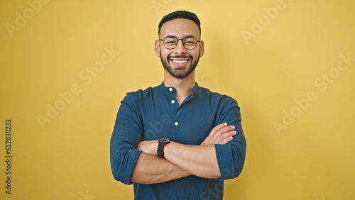 Young hispanic man standing with arms crossed gesture over isolated yellow background